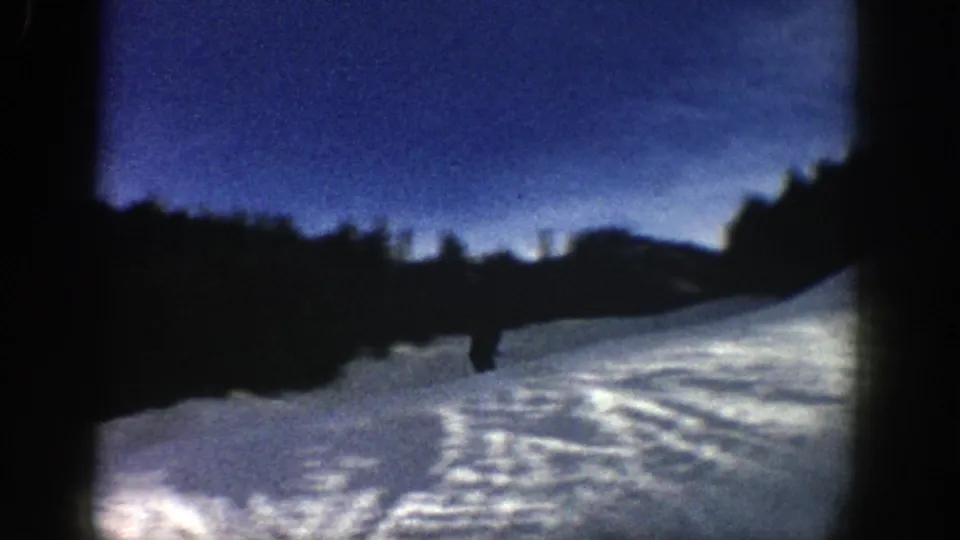 A small skier crosses a shaded Aspen snow slope, with tracked snow and dark hillside shapes in 1961 8mm footage.