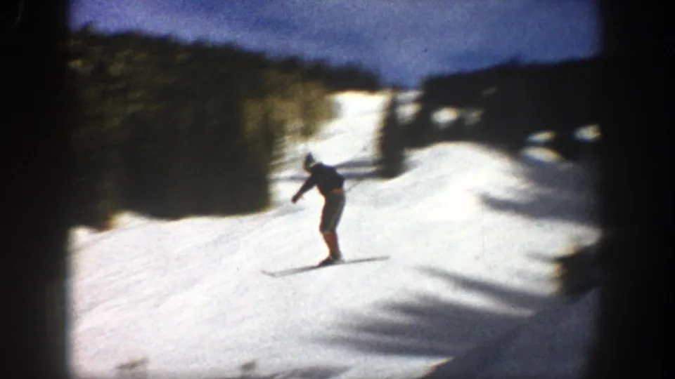 A skier passes close to the handheld camera on a bright Aspen slope, with dark trees and snow shadows visible in 1961 footage.