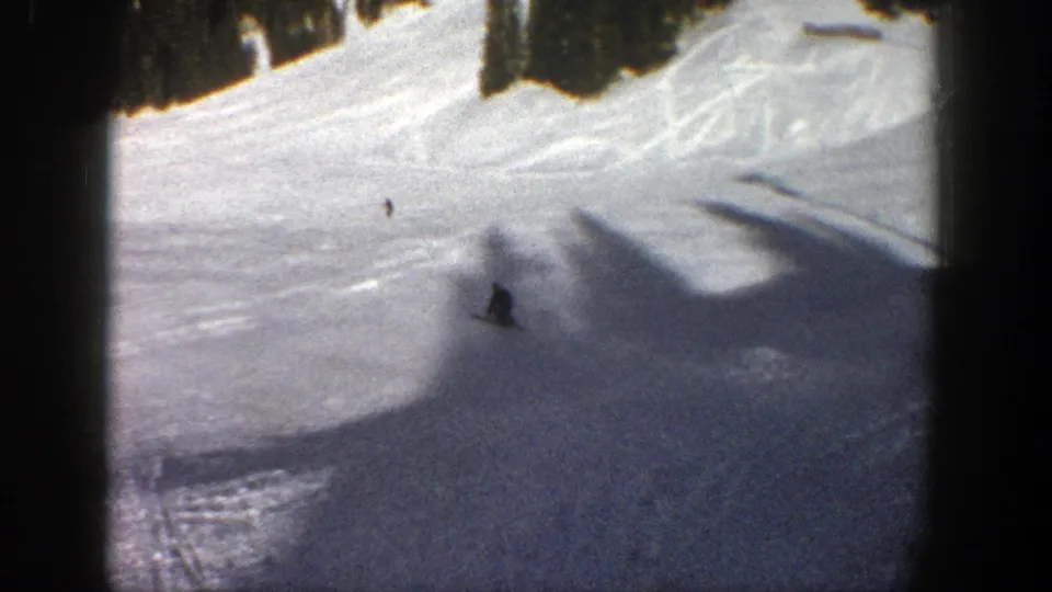 Small skiers move across a bright Aspen ski slope, with dark evergreens framing the upper edge of the 8mm frame.