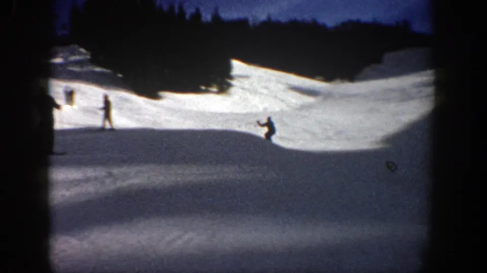 A skier appears low to the snow during or after a jump on an Aspen slope, while another figure stands farther left.