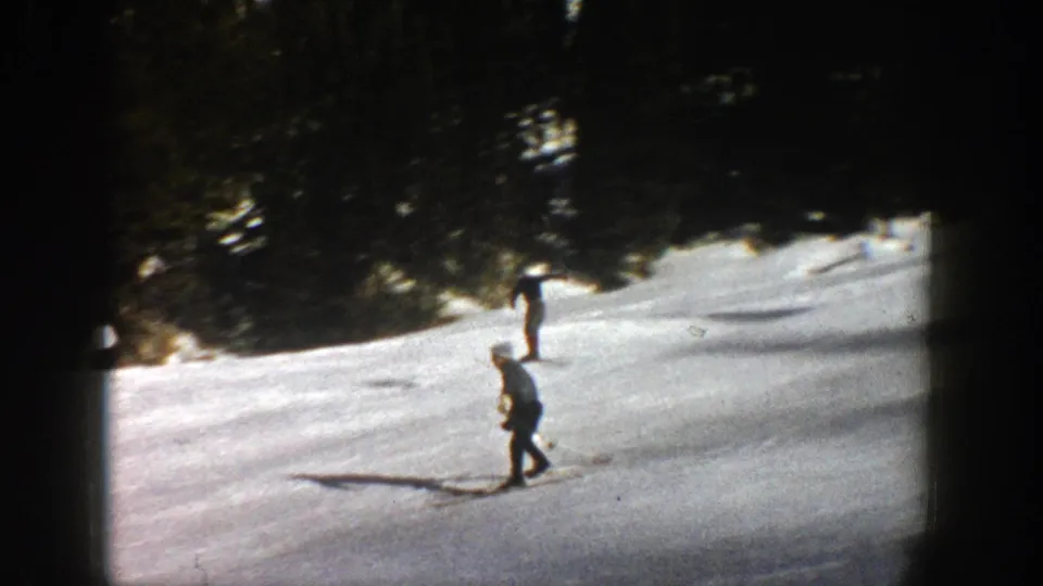 Two skiers are visible at different distances on a bright Aspen snow slope, with dark evergreens along the upper edge.