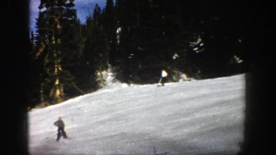 Two skiers appear small against the open snowy hillside in 1961 Aspen, Colorado, with trees and strong sunlight behind them.