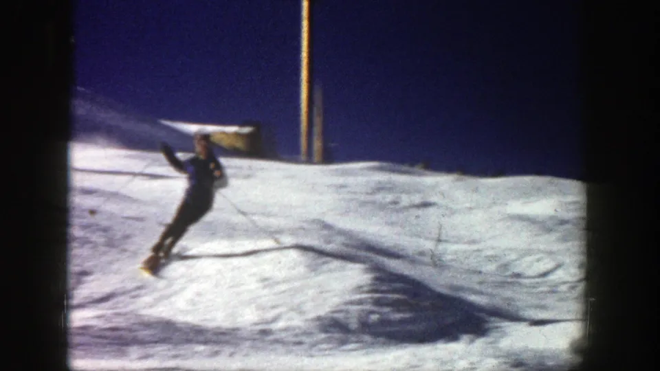 A skier leans into a foreground turn on a 1961 Aspen snow slope, with blue sky and 8mm film borders visible.