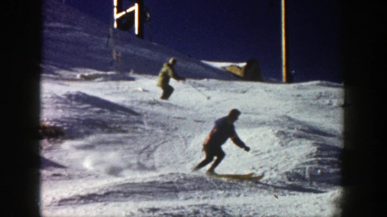 1961 Aspen home-movie frame showing two skiers moving down a bright snow-covered slope near simple resort poles.