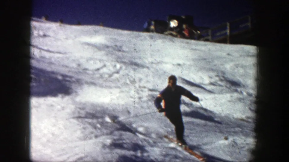 A skier in dark winter clothing crosses the Aspen snow slope, with distant resort fencing and equipment visible above.