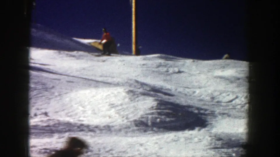 A skier pauses or turns near a snowy rise in Aspen, with a vertical resort pole and dark sky behind.