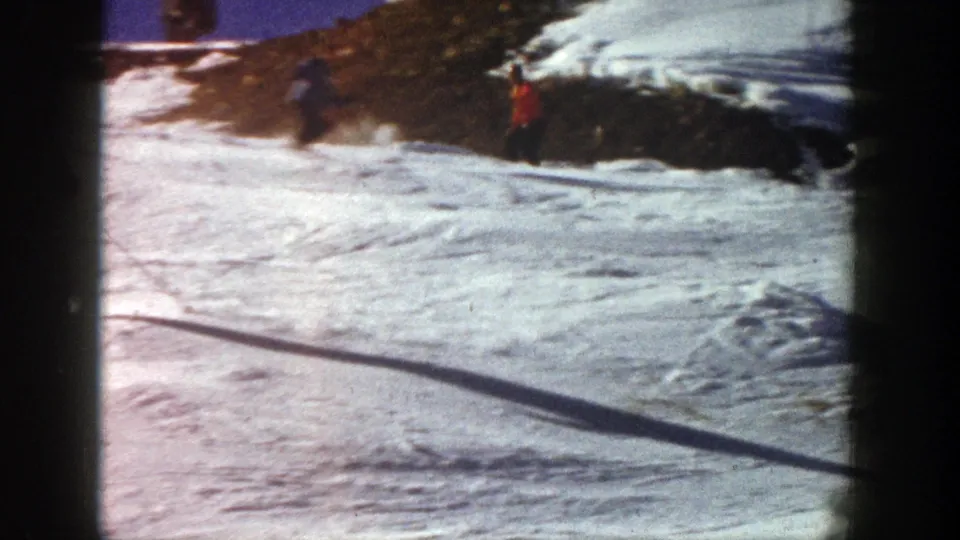 1961 Aspen footage frame with a red-jacketed skier moving along a snow run beside exposed hillside terrain.