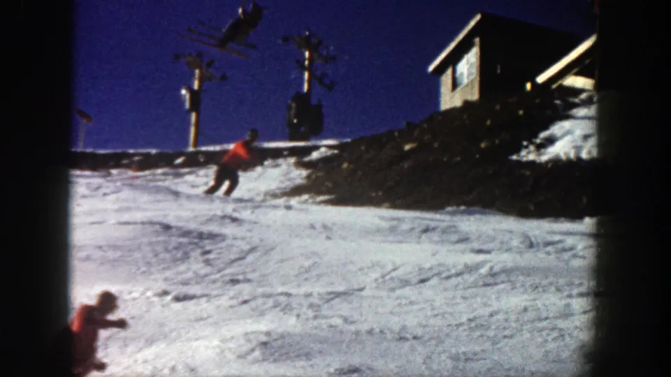 Two skiers descend an Aspen resort slope in 1961, with lift poles and a small building visible above them.
