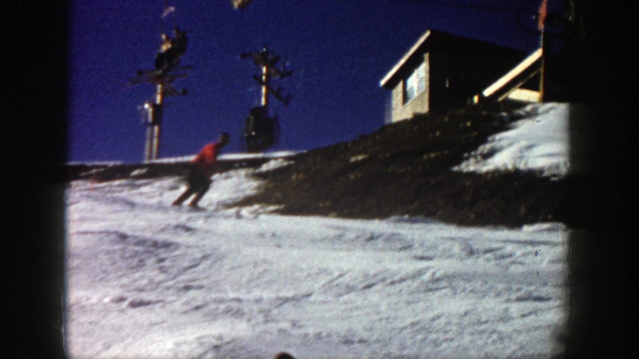 1961 Aspen home-movie frame showing a skier in a red jacket moving downhill on snow below a wooden lift or lodge structure under a dark blue sky.