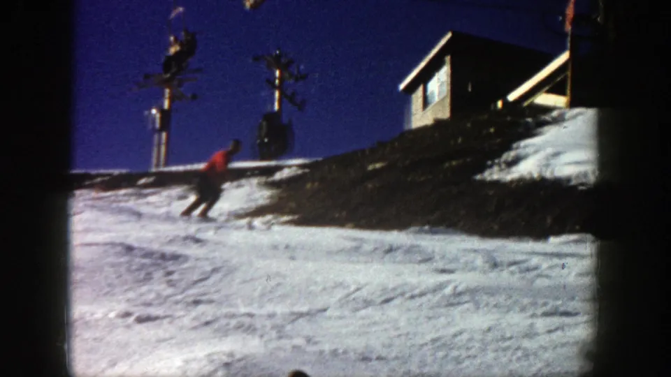 1961 Aspen home-movie frame showing a skier in a red jacket moving downhill on snow below a wooden lift or lodge structure under a dark blue sky.