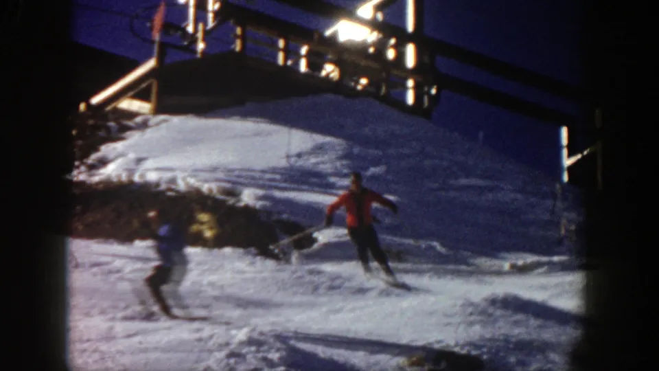 1961 Aspen home-movie frame showing two skiers on a resort slope, with one in red and another darker figure moving through the foreground below the lift-area structure.