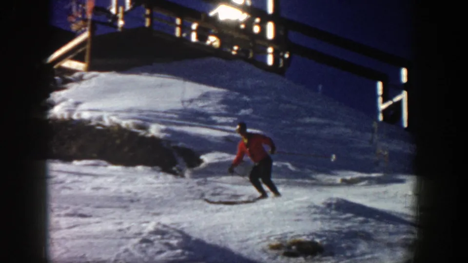 1961 Aspen frame showing a skier in a red jacket turning or gliding downhill beside ski tracks beneath a wooden lift-area platform.