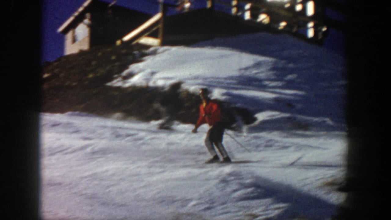 In 1961 Aspen footage, a skier in an orange jacket glides downhill on a snowy slope with a building and lift structure above.