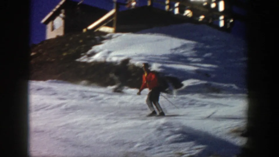 In 1961 Aspen footage, a skier in an orange jacket glides downhill on a snowy slope with a building and lift structure above.