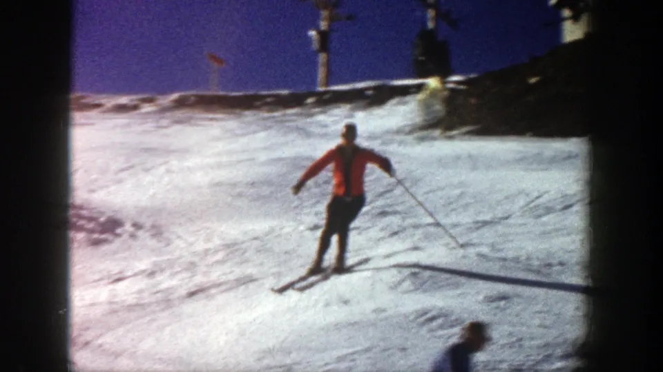 A skier in an orange jacket crosses the Aspen snowfield, with poles extended and lift equipment visible near the upper ridge.