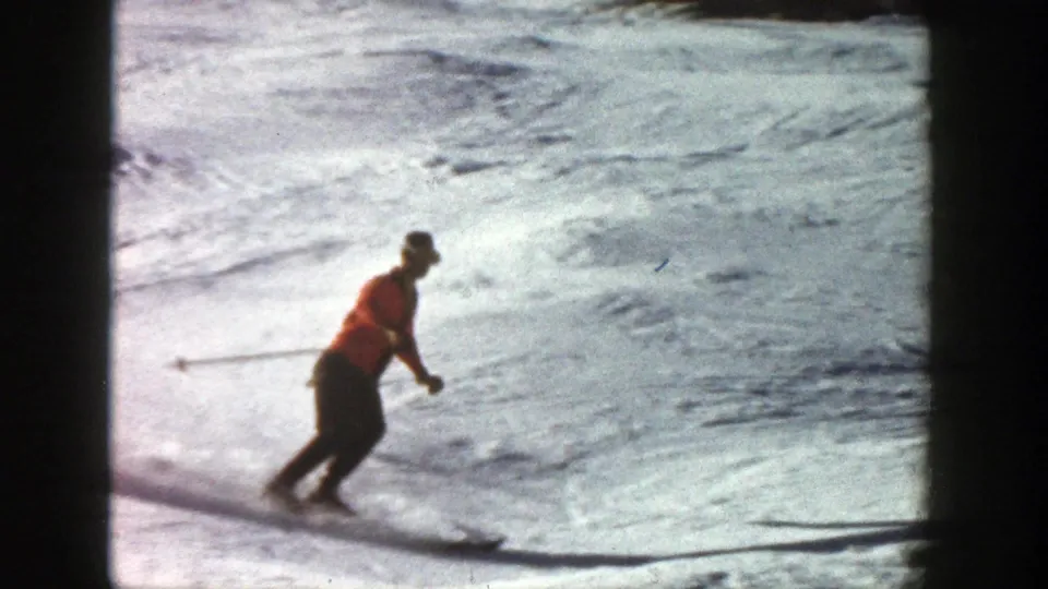 The orange-jacketed skier leans through a turn on the Aspen slope, seen in soft 8mm color with bright snow and shadowed tracks.