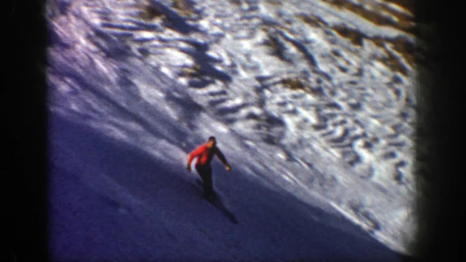 The red-jacketed skier glides along a shadowed Aspen slope with textured snow and tracks visible nearby.