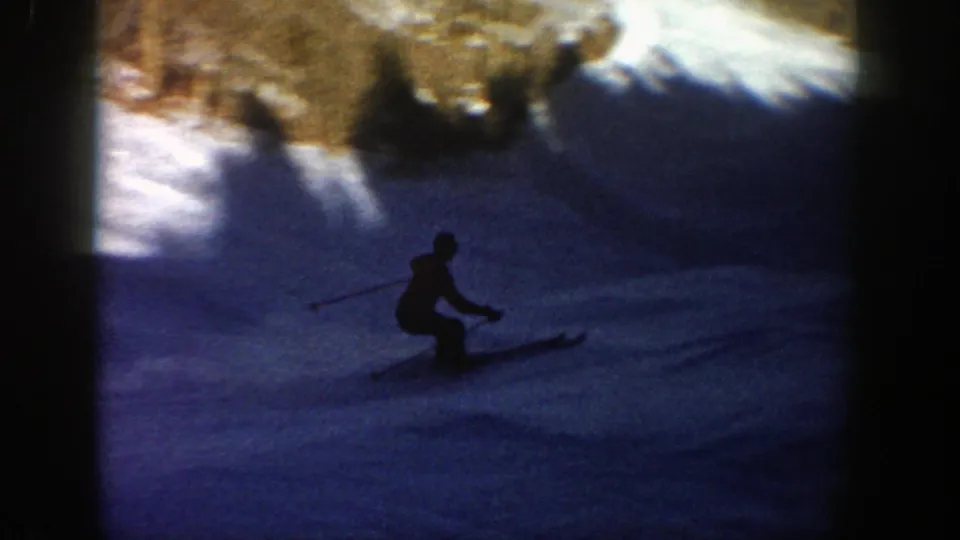 A skier appears mostly in silhouette on a shaded Aspen snow slope, with ski poles and bright snow above the shadow line.