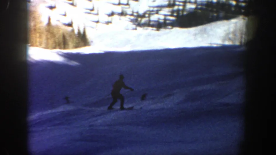 A distant skier continues down a broad shaded slope in the 1961 Aspen home-movie footage, with trees and bright snow in the background.