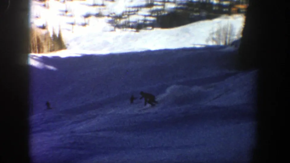 Near the end of the Aspen ski clip, the skier bends low while crossing a shaded snowfield with bright mountain snow beyond.