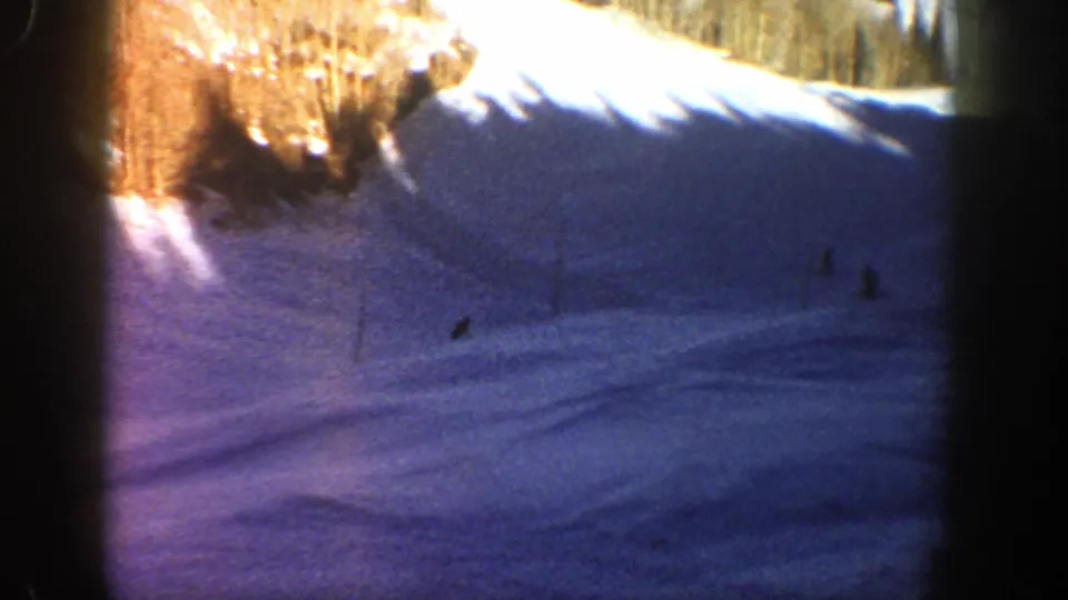 Wide 1961 Aspen frame with several small riders or skiers on a snow-covered slope under a sunlit tree line.