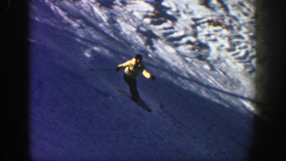 A skier in 1961 Aspen footage descends a snowy slope with poles, framed by dark 8mm borders and blue winter shadows.