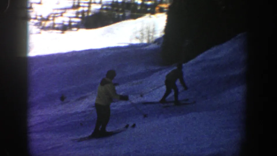 Two skiers cross a blue-shadowed Aspen slope in 1961 home-movie footage, with trees or slope edges visible in the bright background.