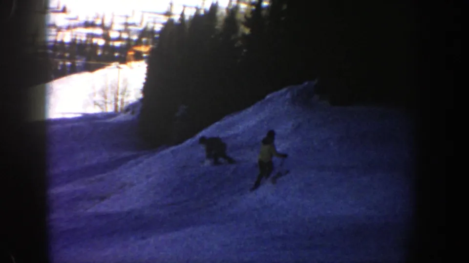 On a snowy Aspen slope, one adult skier is down or losing balance while another skier continues downhill in the 1961 home movie.