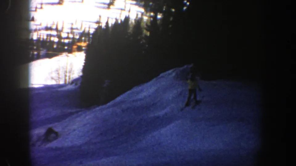 The 1961 Aspen footage shows one skier continuing downhill as a second figure remains low on the snow near the edge of the frame.