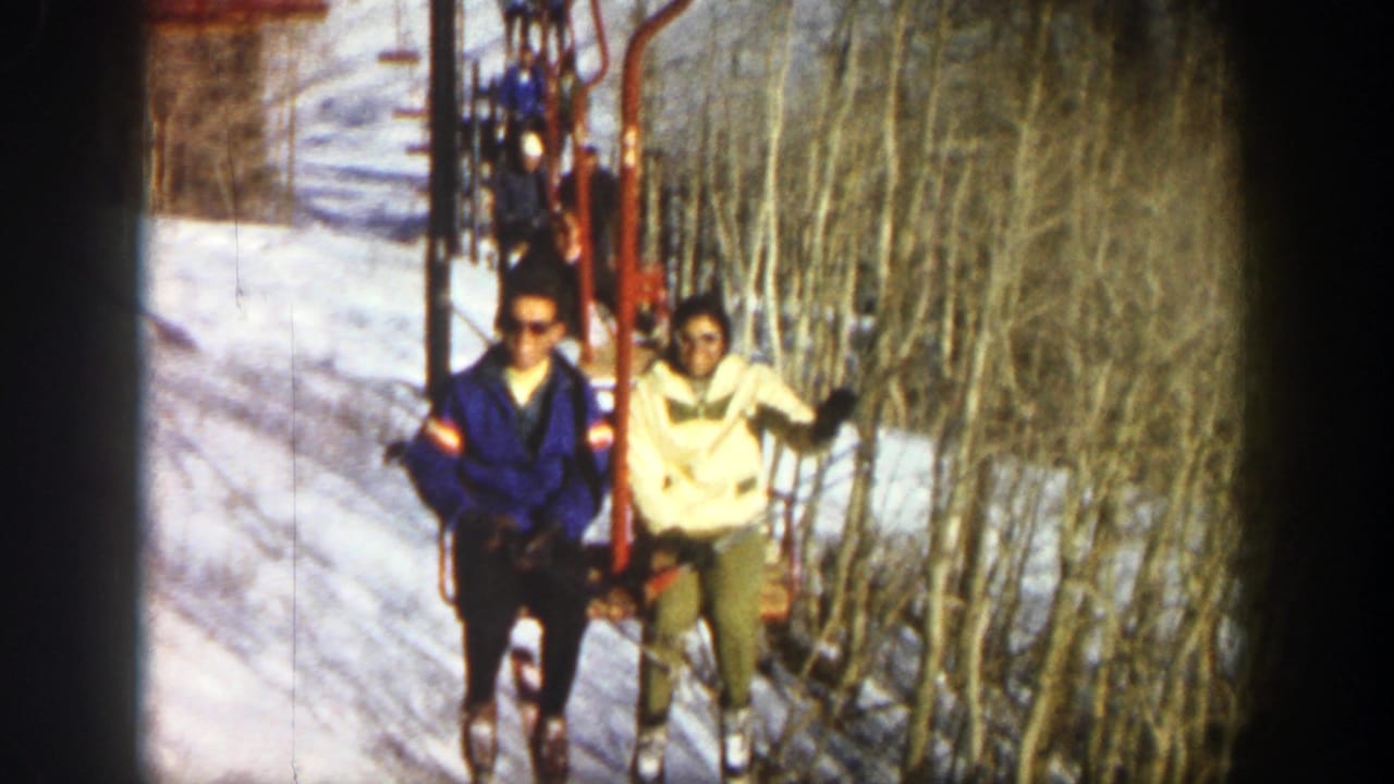 In 1961 Aspen, two skiers ride a chairlift, one in a blue jacket and sunglasses and one in a cream jacket beside bare trees.