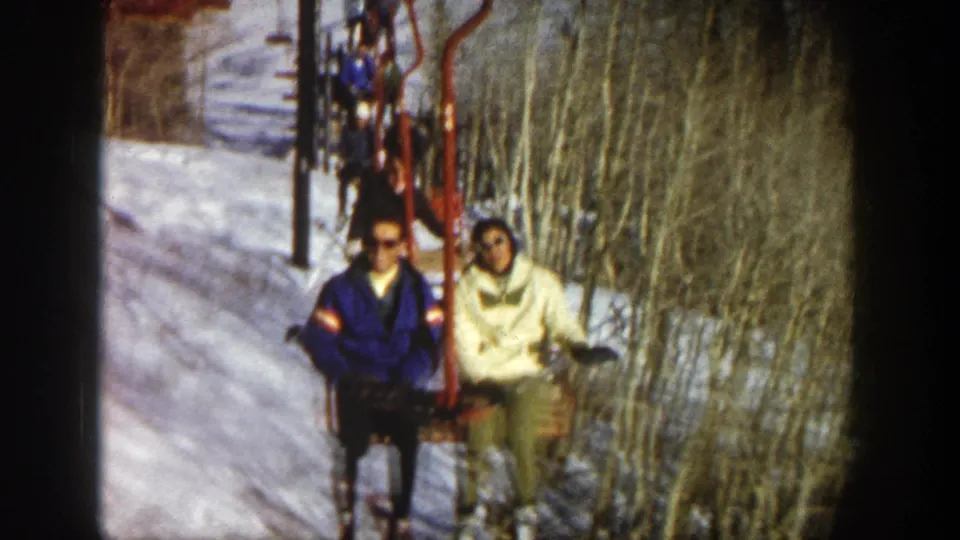 Two skiers on a red chairlift ascend through a snowy Aspen slope in 1961, with another chair and bare trees behind them.