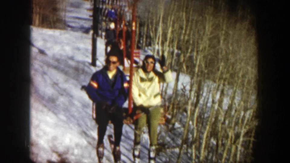 On a 1961 Aspen chairlift, the skier in a cream jacket raises a hand while sitting beside a skier in a blue jacket.