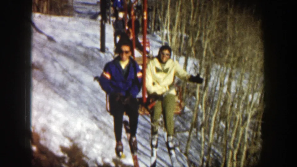 The two Aspen skiers pass near the camera on the chairlift, with skis dangling above snow and bare trees beside the lift line.
