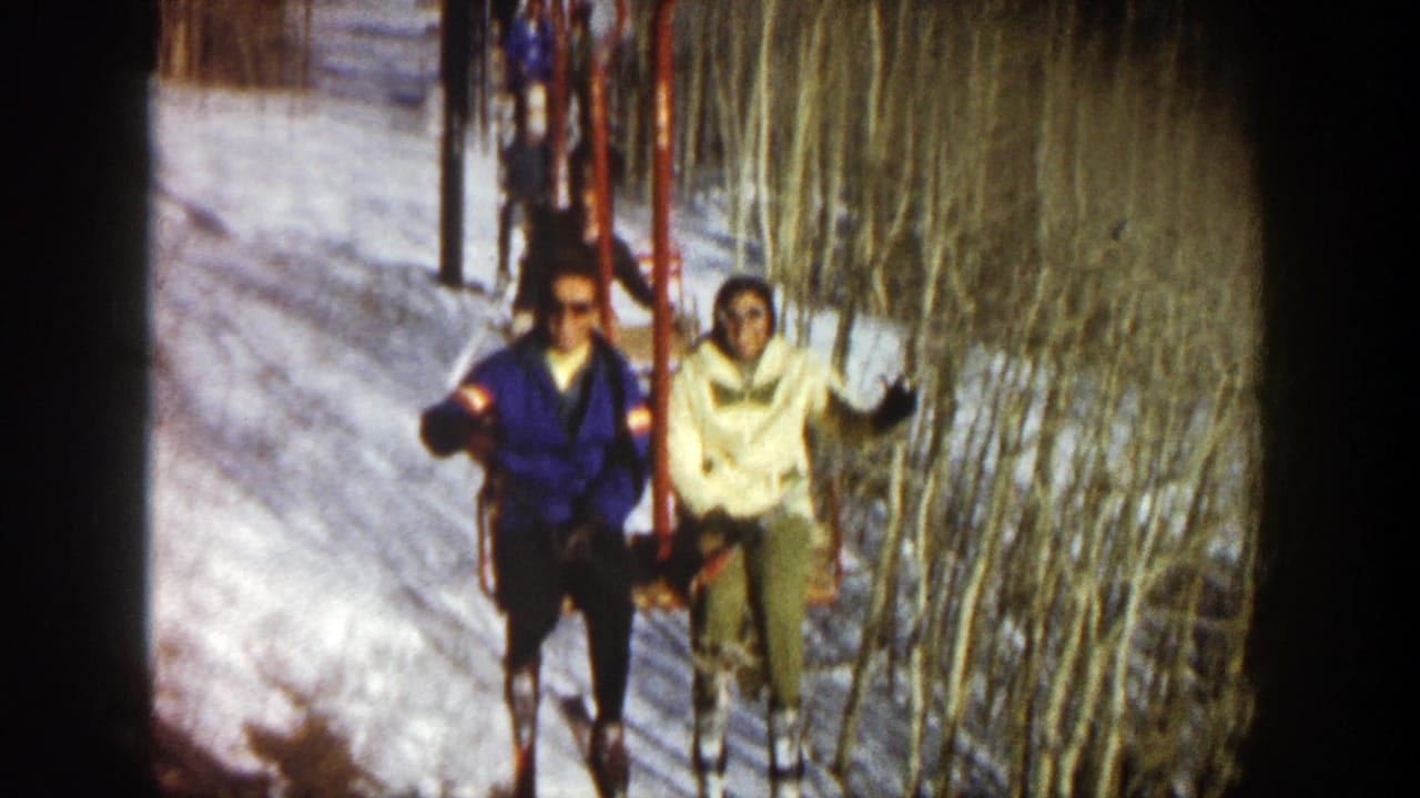 Two riders on a red chairlift wave above a snowy slope lined with bare trees.