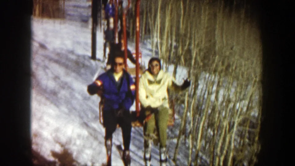 In 1961 Aspen, Colorado, two chairlift riders in blue and cream jackets wave toward the camera above snow and bare aspen trees.