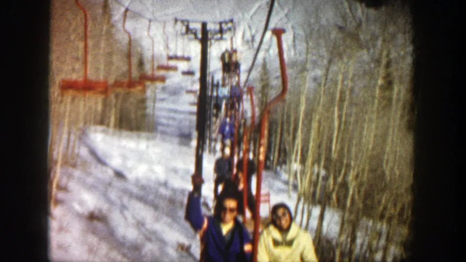 Two visible riders wave from a red chairlift in the Aspen ski area, with snow, lift poles, cables, and bare trees behind them.