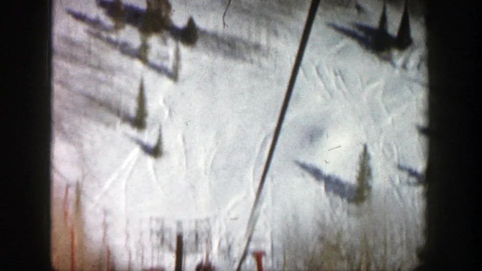 A high, soft 8mm view looks down over snow, chairlift shadows, poles, and bare trees during a 1961 Aspen ski outing.