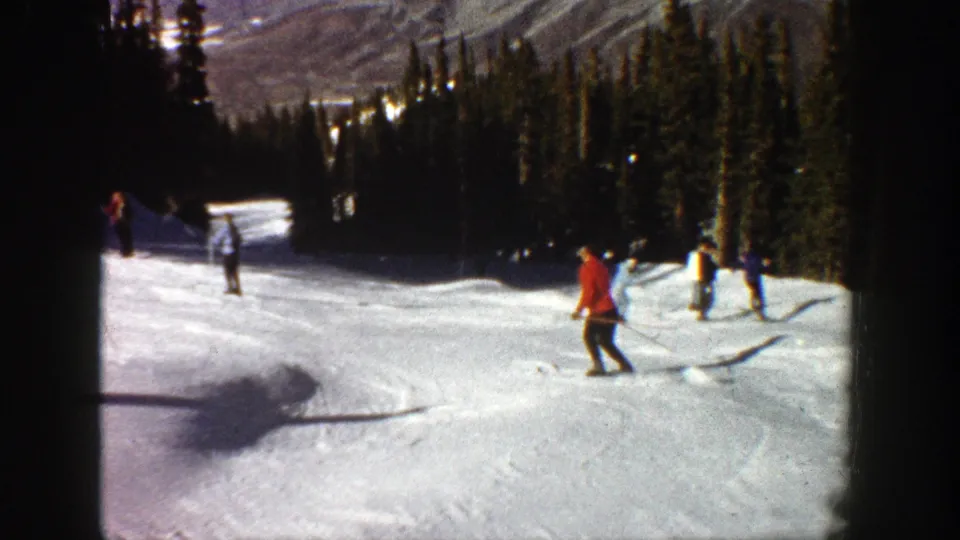 Skiers move across an Aspen slope in 1961, with evergreen trees, mountain terrain, and bright snow visible in the home-movie frame.