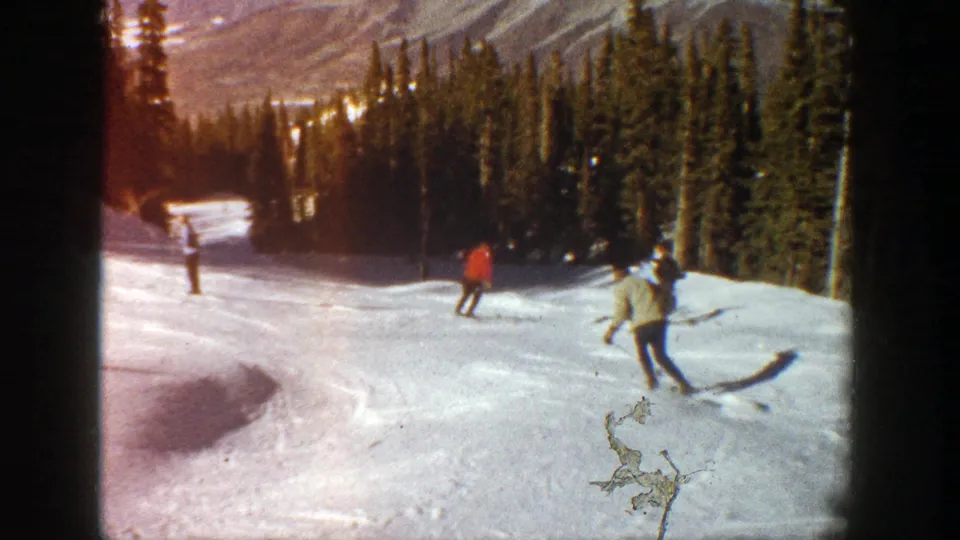 1961 Aspen skiing footage with a red-coated skier and another skier crossing a bright run near evergreen trees.