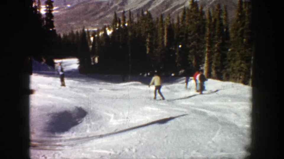 1961 Aspen home-movie view of skiers descending a groomed-looking snowy slope with evergreens and mountains beyond.