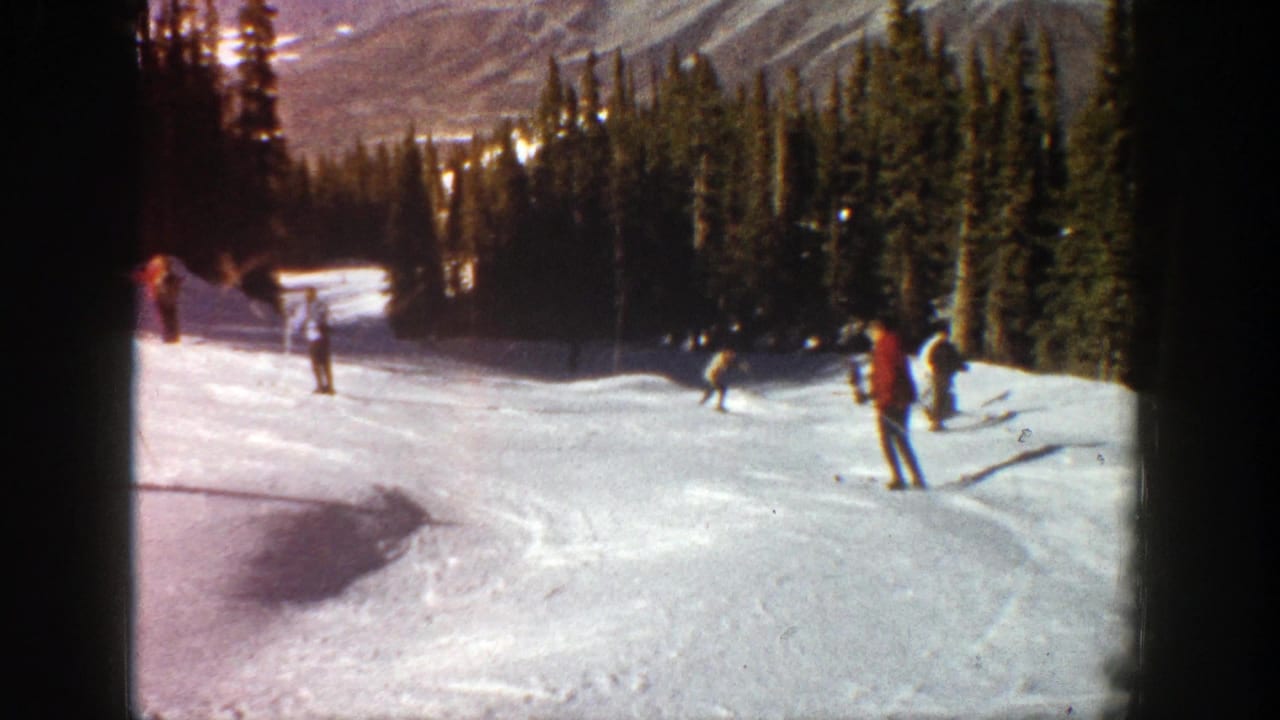 1961 Aspen home-movie frame showing several skiers on a snowy mountain run with evergreens and distant slopes behind them.
