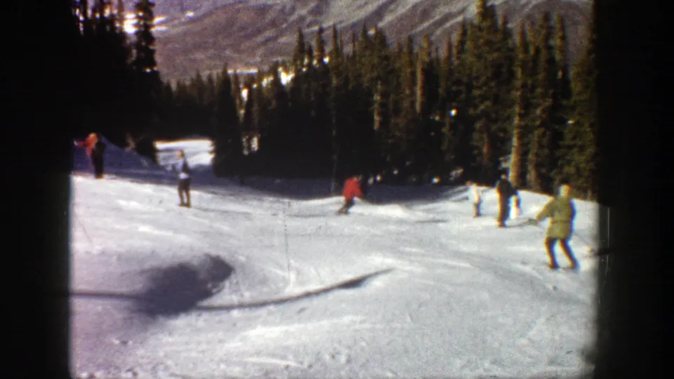 1961 Aspen home-movie frame showing skiers in red, light, and dark coats moving across a tree-lined snowy run.