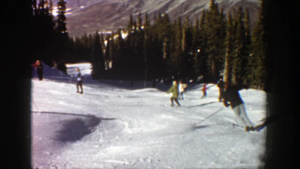 1961 Aspen frame with a skier at the right edge and several distant skiers on a snowy run beside dense evergreens.