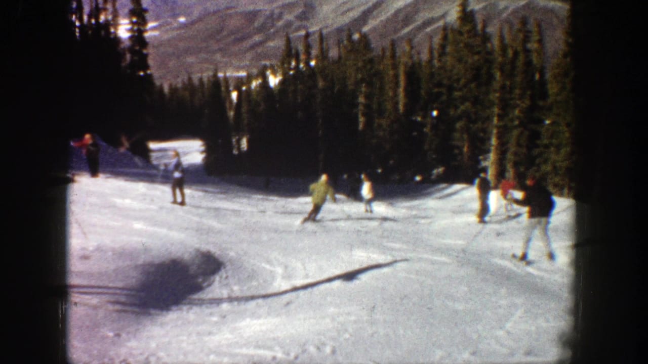 1961 Aspen home-movie frame showing several skiers on a sunlit snow run with evergreen trees and mountain ridges behind them.