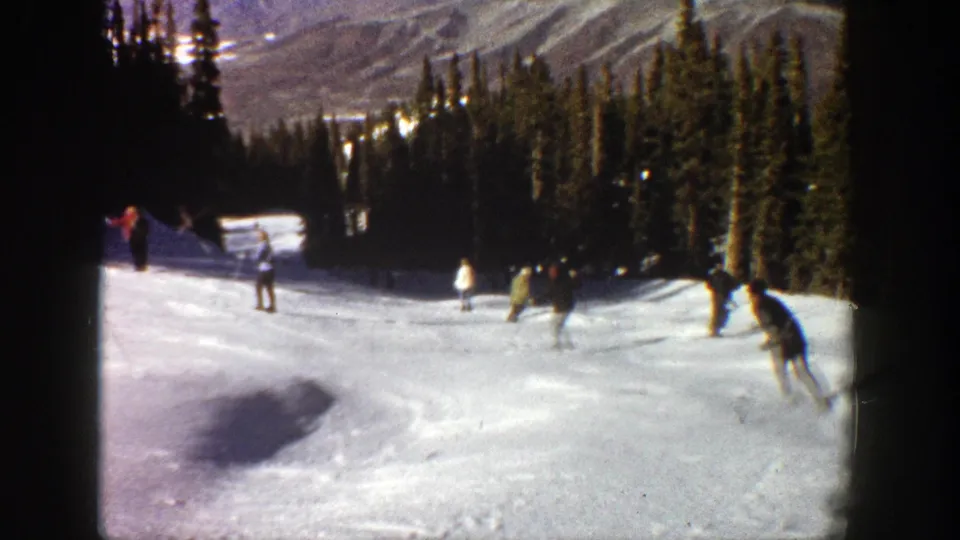 1961 Aspen ski footage frame with a skier near the right edge and other figures spread across a bright run beneath dark evergreens.