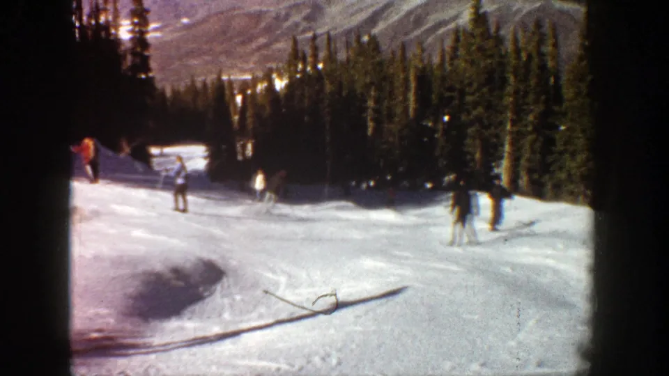 1961 Aspen home-movie frame showing skiers spaced across a snowy trail, with dark evergreen trees and hazy mountain ridges beyond.