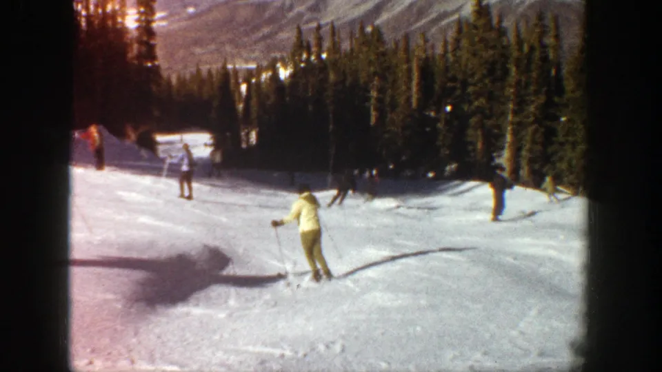 1961 Aspen frame with a light-coated skier in the foreground and other skiers farther up the snow run near evergreen trees.