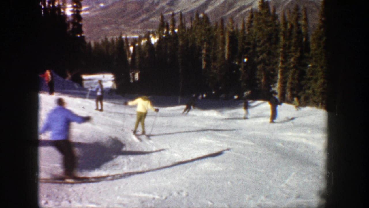 1961 Aspen home-movie frame showing several skiers on a sunlit snowy run with evergreens and mountains behind them.