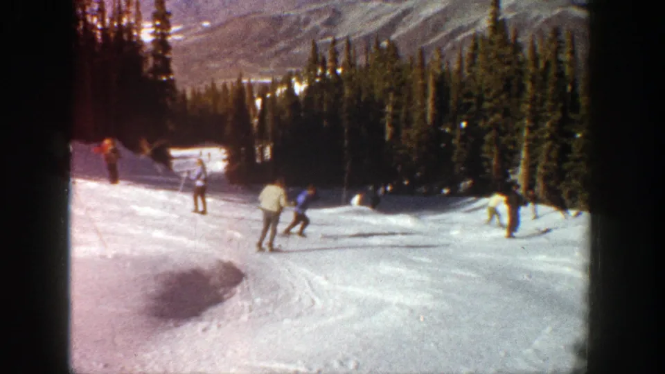 1961 Aspen 8mm frame showing skiers spaced across a downhill run, with evergreen trees and mountain scenery beyond.