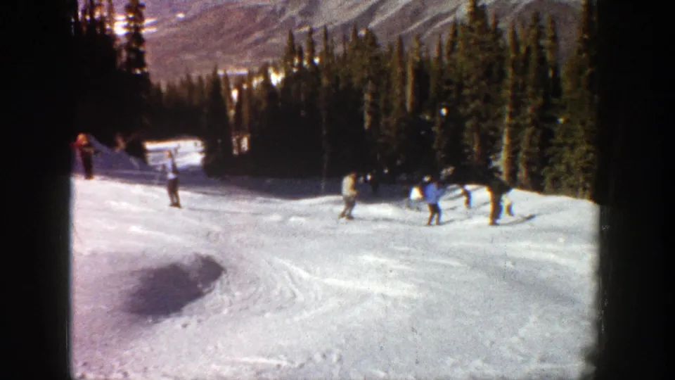 1961 Aspen home-movie frame with skiers on a sunlit snow run near dense evergreen trees and mountain terrain.